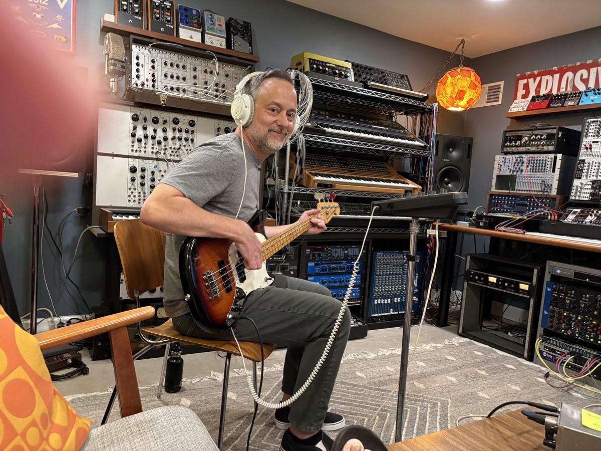 The bass player smiling in headphones, surrounded by synthesizers and speakers