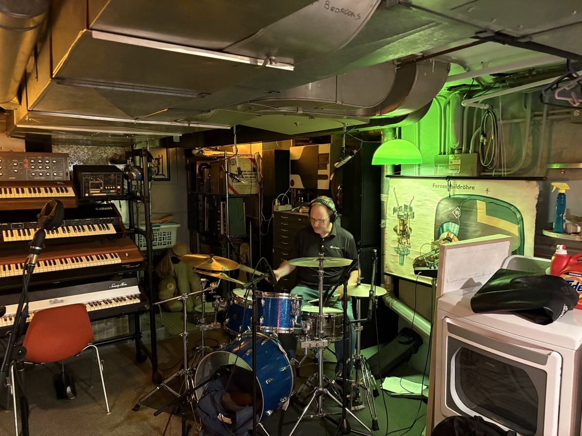 The drummer in headphones at his kit in a basement studio lined with vintage keyboards