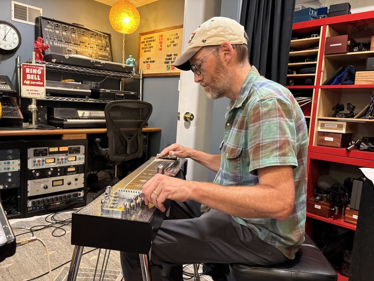 The pedal steel player working the bar, with studio gear and a clock on the wall behind him